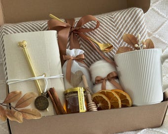 May include: A gift box containing a white mug, a notebook, a pen, a blanket, a jar of honey, a honey dipper, dried orange slices, and a small bag tied with a ribbon. The box is brown and the items are arranged inside.