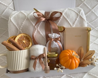 May include: A gift basket featuring a white waffle-weave blanket, a mug with cinnamon sticks and dried orange slices, a small pumpkin, a jar of honey, and a notebook with the word "NOTES". The basket is tied with a brown ribbon.