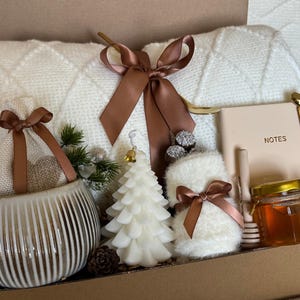 May include: A gift box containing a white blanket, mug, candle, notepad, honey, and other items. The mug and candle are white, and the blanket has a diamond pattern. The box is tied with brown ribbons and includes a small pine tree branch.