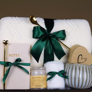 May include: A white knitted throw blanket, a green ribbon, a candle, a mug, a notebook with a gold pen, a heart-shaped box, and a white fluffy item in a gift basket.