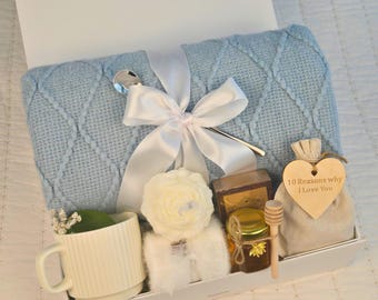 May include: A gift basket filled with a blue knitted throw blanket, a white mug, a white soap, a jar of honey, a wooden honey dipper, a small white fluffy towel, and a small burlap bag with a wooden heart that says "10 Reasons Why I Love You".