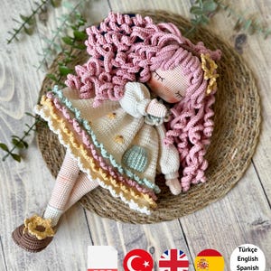 May include: A crochet doll with long curly pink hair, wearing a cream-colored dress with a light green pocket and a multi-layered ruffled hem in yellow, light blue, and white. The doll has brown shoes with yellow flower accents and a PDF pattern label.