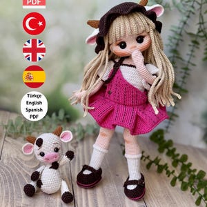 Op de afbeelding: Een handgemaakte gehaakte pop met lang blond haar, gekleed in een roze jurk en bruine schoenen. Een kleinere gehaakte koeienpop staat naast de grotere pop. De tekst luidt "PDF PATTERN ONLY - NOT FINISHED DOLL."