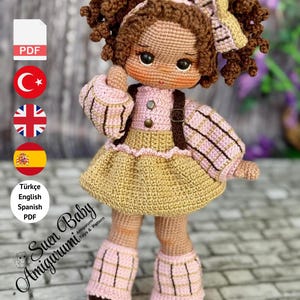 May include: A handmade crochet doll with brown curly hair, wearing a pink and tan dress, shoes, and a headband. The doll has a backpack and is standing. The image includes text that says "Suen Baby Amigurumi" and "PDF PATTERN ONLY - NOT FINISHED DOLL."
