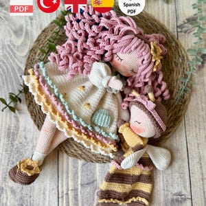 May include: A crochet pattern PDF featuring a large doll with curly pink hair and a cream dress, alongside a smaller bee-themed doll with yellow and brown stripes. The text reads: PDF, Türkçe, English, Spanish, PDF, PDF PATTERN ONLY – NOT FINISHED DOLL.