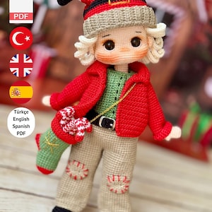 以下が含まれることがあります： 赤いジャケット、グリーンのシャツ、ベージュのズボンを着た手作りの編み人形。人形はサンタ風の帽子をかぶり、小さな靴下を持っています。「PDF PATTERN ONLY - NOT FINISHED DOLL」というテキストが表示されています。