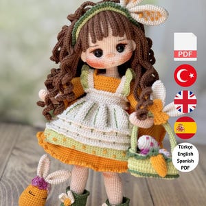 Puede incluir: Muñeca de ganchillo hecha a mano con cabello rizado castaño, vestida con un vestido naranja y un delantal blanco. La muñeca tiene una diadema de orejas de conejo y sostiene una cesta con huevos de Pascua. Un conejo de ganchillo está al lado de la muñeca.