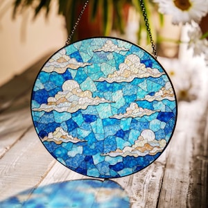 May include: A round stained glass window depicting a blue sky with white clouds. The glass is cut into small pieces and arranged in a mosaic pattern. The window is framed in a black metal frame and is hanging from a chain.
