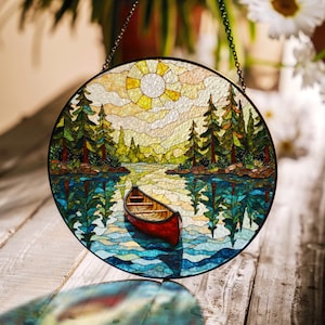 Pode incluir: Painel redondo de vitral retratando uma cena de lago com uma canoa vermelha, árvores e um sol. A obra de arte é emoldurada com uma borda de metal preto e pendurada por uma corrente. A água reflete as árvores e o céu.