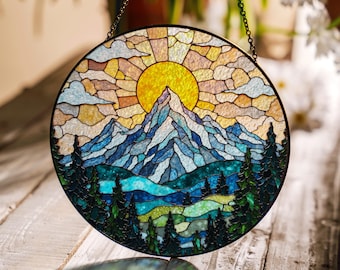 May include: Round stained glass panel depicting a mountain landscape with a yellow sun, blue mountains, and green trees. The panel is framed in black and hangs from a chain. The design is reminiscent of a sunrise over a mountain range.