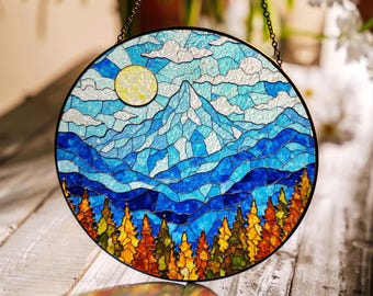 Può includere: Un acchiappasole in vetro colorato che raffigura una catena montuosa con un sole giallo e un cielo blu. Le montagne sono blu e bianche, e gli alberi sono arancioni, gialli e verdi.