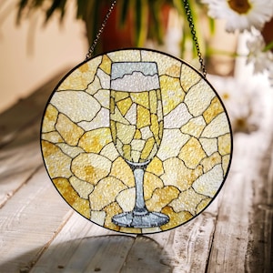 Puede incluir: Una ventana redonda de vidrio emplomado colgada de una cadena. La ventana presenta un diseño de vidrio amarillo y blanco de una copa de champán.