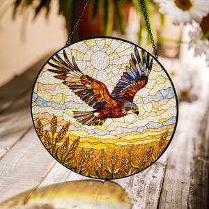 May include: A round stained glass window featuring a soaring hawk with outstretched wings. The hawk is depicted in shades of brown, orange, and blue against a backdrop of a sun and wheat field. The window is framed in black and hangs from a chain.