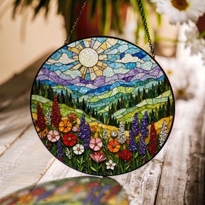 Op de afbeelding: Een gebrandschilderd zonnenvanger met een kleurrijk landschap met een heldere zon, blauwe lucht, groene heuvels en een bloemenveld. De zonnenvanger is rond en heeft een metalen ketting om op te hangen.
