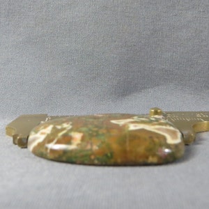 May include: A close-up of a brown, green, and white agate cabochon with a polished surface. The cabochon is mounted in a brass setting with a ruler.