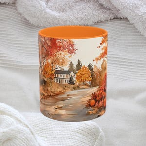 May include: A ceramic coffee mug with an orange interior and a watercolor painting of a fall landscape. The artwork depicts a road leading to a white house, surrounded by trees with red, orange, and yellow leaves.