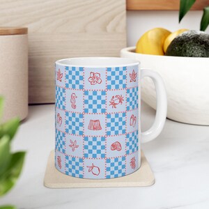 May include: White ceramic mug with a blue and white checkered pattern. The pattern includes red illustrations of a seahorse, starfish, hibiscus flower, crab, shorts, seashell, and other beach-themed icons. The mug is on a beige coaster.