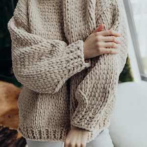 Puede incluir: Un suéter de punto grueso beige de corte holgado y mangas largas.