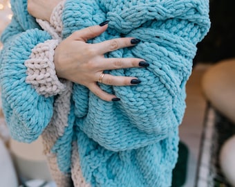 May include: A blue and cream chunky knit cardigan with a cable knit pattern. The cardigan is open at the front and has a relaxed fit.