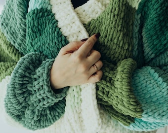 May include: A chunky knit cardigan with a white, green and blue colour block pattern. The cardigan has a v-neck and a button closure.