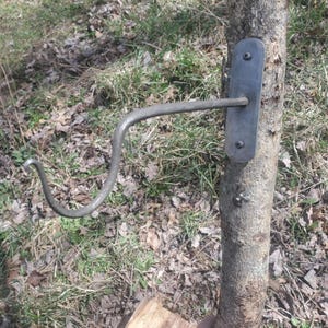 Puede incluir: Un gancho de metal sujeto a un tronco de árbol con una placa rectangular de metal. El gancho es curvo y tiene un extremo puntiagudo.