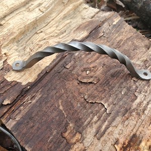 May include: A hand-forged, twisted metal handle with a matte gray finish. The handle has a curved design with a spiral pattern and mounting holes on each end. The hardware is displayed on a weathered wooden surface.