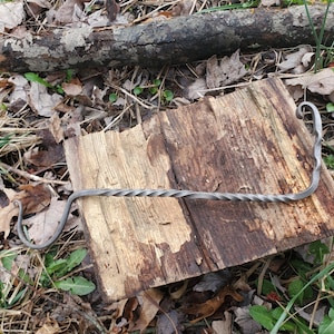 Peut inclure: Un tisonnier en métal forgé à la main, avec une tige torsadée et des extrémités incurvées. Le tisonnier gris foncé repose sur une surface en bois vieilli, entouré de feuilles et d'une bûche. Le design suggère une esthétique rustique et artisanale.