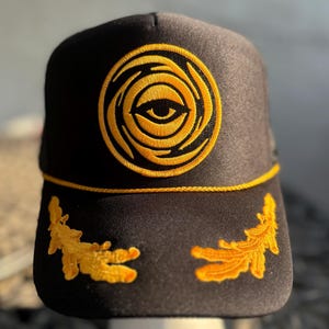 Puede incluir: Gorra de béisbol negra con un diseño de ojo bordado en dorado en la parte delantera. La gorra tiene ribetes dorados y diseños de hojas doradas en los laterales. La gorra se exhibe sobre un soporte blanco.