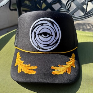 Puede incluir: Gorra de camionero negra con un diseño de ojo blanco y negro en el centro. La gorra tiene un borde bordado amarillo y dos diseños de hojas amarillas en la parte delantera. La parte trasera de la gorra es de malla negra.