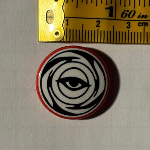May include: A black and white button with a red border. The button features a stylized eye with a swirling pattern around it.