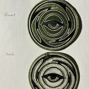 Puede incluir: Vista frontal y posterior de un parche bordado verde y negro con un diseño de ojo estilizado. El ojo es verde con una pupila negra y reflejos blancos. El parche es circular y tiene un borde blanco.
