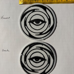 Puede incluir: Dos parches bordados blancos con un diseño de ojo negro. El ojo está rodeado por un patrón de remolino. El texto "Front" y "Back" está escrito en la imagen.