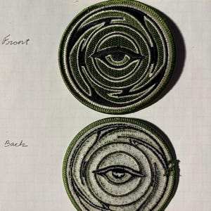 Puede incluir: Un par de parches redondos bordados de color verde con un diseño de ojo en blanco y negro. La parte delantera del parche tiene un ojo verde, mientras que la parte trasera del parche tiene un ojo blanco.