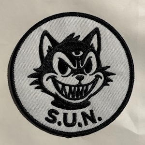 Op de afbeelding: Geborduurde patch met een zwarte kat met een grijns en een derde oog, op een witte achtergrond binnen een zwarte cirkel. De tekst "S.U.N." staat onder de kat.