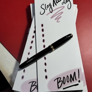 May include: A stack of white notepads with a pink and black design. The top notepad has the text "Slay All Day" written in black cursive. The bottom notepad has the text "BOOM!" written in black cursive. A black pen is lying on top of the notepads.