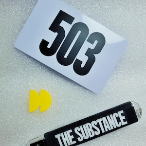 May include: A white card with the number 503 printed in black. A yellow plastic object shaped like a Pac-Man character. A black USB drive with the text "THE SUBSTANCE" printed in white.