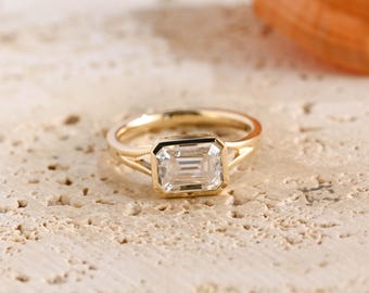 May include: A gold ring featuring a clear, rectangular emerald-cut gemstone set in a bezel setting. The band is thin and smooth, with a split shank design that connects to the central stone setting, showcasing a polished metallic finish.