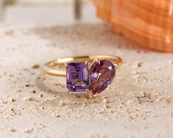 May include: A gold ring featuring two distinct gemstones: a rectangular emerald-cut purple amethyst and a pear-shaped, multi-toned purple and reddish-pink gemstone. The stones are set side-by-side on a thin, polished gold band with four-prong settings.