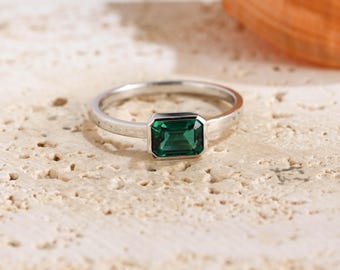 May include: A silver-toned ring featuring a single emerald-green rectangular gemstone set in a bezel setting. The band is smooth and polished, showcasing a clean, minimalist design that highlights the vibrant green color of the central stone.