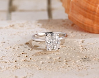 May include: A silver-toned ring featuring a single cushion-cut clear gemstone set in a four-prong setting. The band is smooth and polished, showcasing a reflective metallic finish that catches the light, highlighting the faceted stone.
