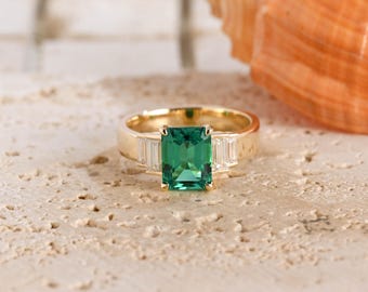 May include: A gold ring featuring a central emerald-cut green gemstone flanked by two smaller baguette-cut clear stones on each side. The band is polished gold, and the setting holds the stones securely in a classic three-stone arrangement.