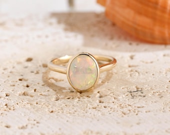 May include: A gold ring featuring a smooth, oval-shaped opal gemstone set in a simple bezel setting. The opal displays iridescent flashes of color, including soft pinks, greens, and yellows, set against a polished gold band that encircles the finger.