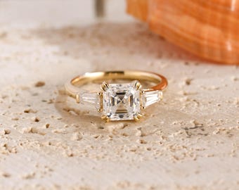 May include: A gold ring featuring a central square-cut stone flanked by two smaller baguette-cut stones on each side. The band is polished gold, and the setting holds the clear, faceted stones securely in place with multiple metal prongs.