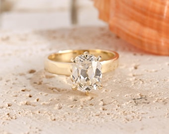 May include: A gold ring featuring a single cushion-cut clear gemstone set in a prong setting. The band is smooth and polished, showcasing a classic design with a central stone that reflects light through its multifaceted surface.