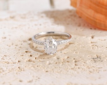 May include: A silver-toned metal ring featuring a central oval-cut clear gemstone. The band is slender and adorned with a series of small, round, clear stones embedded along the shank on both sides of the prominent oval center stone.