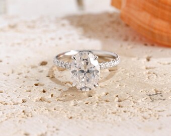 May include: A silver-toned ring featuring a large, oval-cut clear center stone held by four prongs. The band is thin and encrusted with a row of small, round, clear pavé-set stones that extend along the sides of the ring shank.
