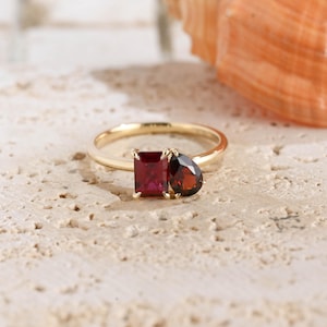 May include: A gold ring featuring two distinct red gemstones set side by side. One stone is a rectangular cut, whilst the other is a pear-shaped cut. The band is a simple, polished gold metal, and the stones are held in place by small gold prongs.
