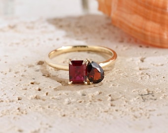 May include: A gold ring featuring two distinct red gemstones set side by side. One stone is a rectangular cut, while the other is a pear-shaped cut. The band is a simple, polished gold metal, and the stones are held in place by small gold prongs.