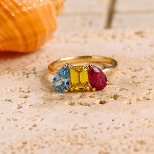 May include: A gold ring featuring three gemstones: a blue teardrop-shaped stone, a yellow rectangular stone, and a red teardrop-shaped stone.
