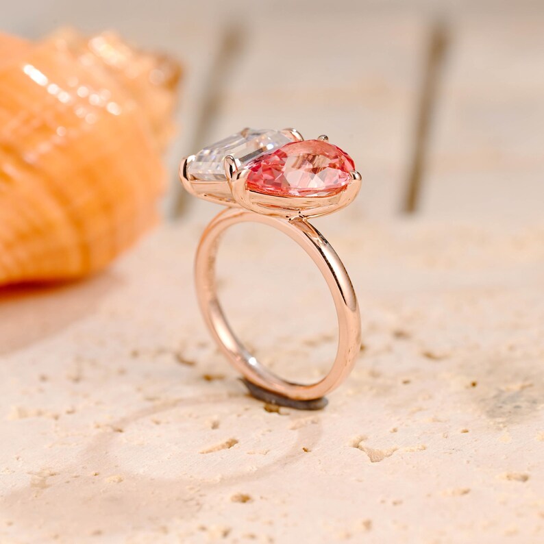 May include: A rose gold ring with a pink and clear gemstone setting. The ring is set on a light brown textured surface.